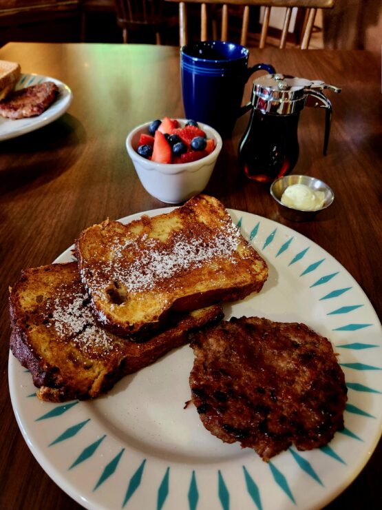 French Toast and Apple Sausage on a breakfast plate, with fresh fruit in a bowl.