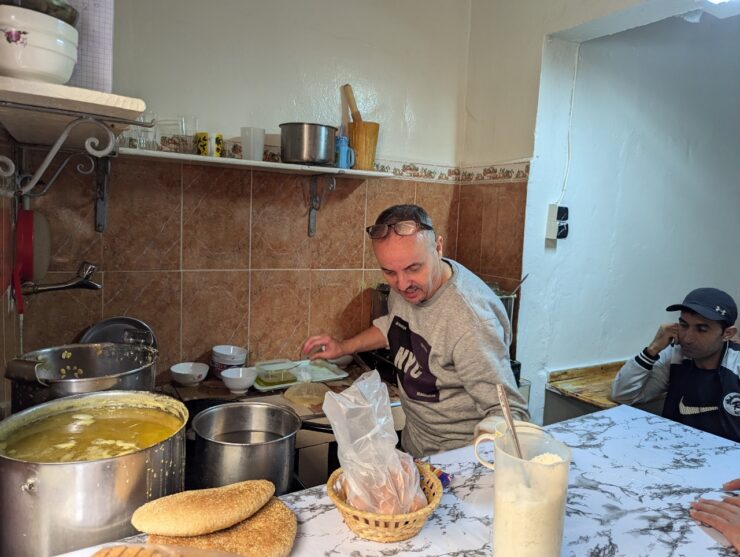 A Moroccan man standing over big pots of woup in a small kitchen. 