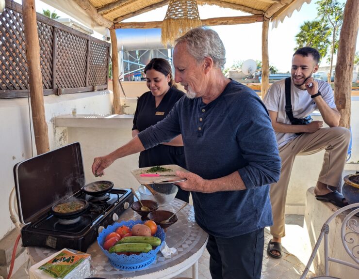 A man sprinkles herbs into tagine stew while the cooking instructor and guide look on. 