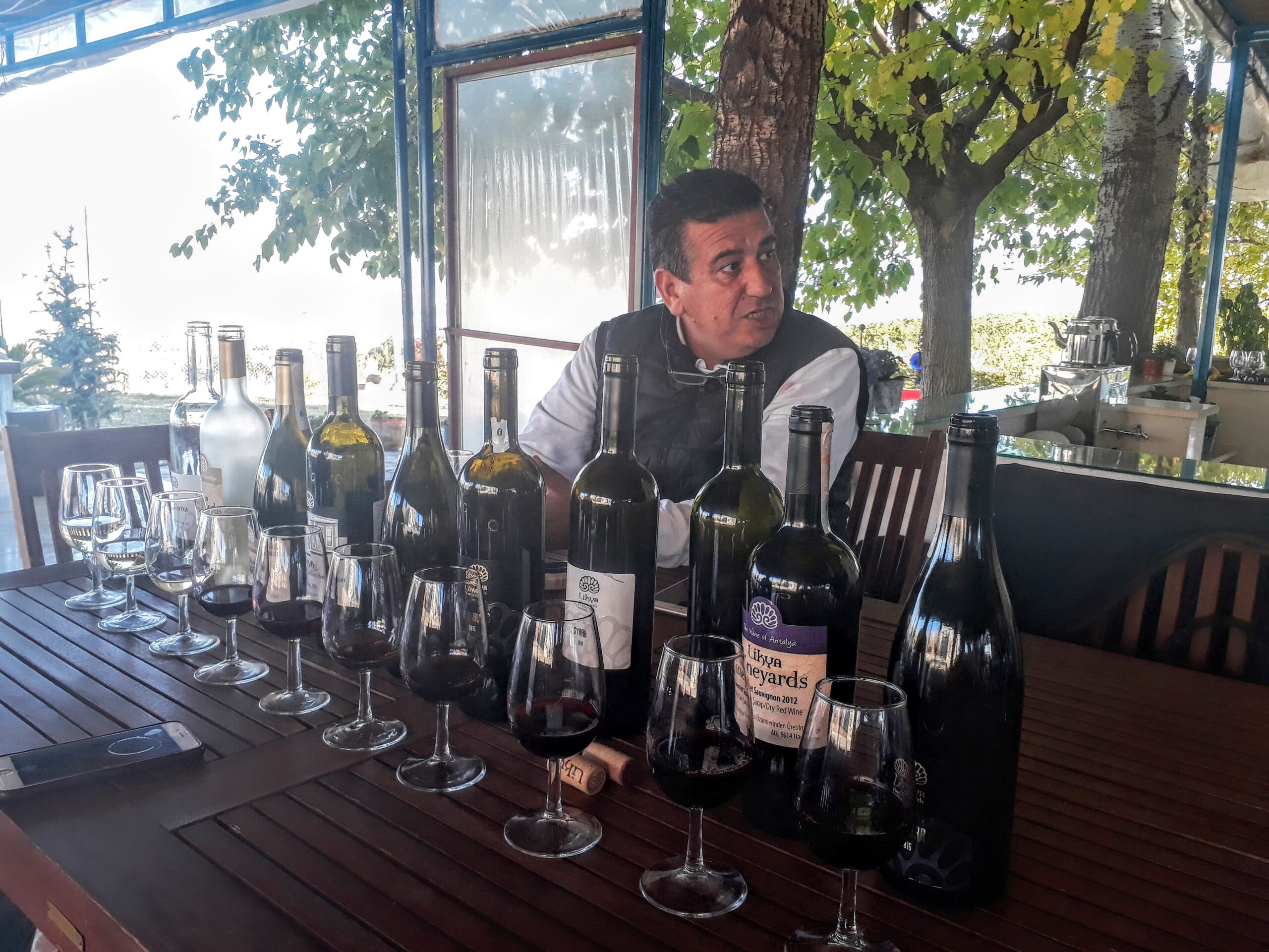 smiling man sitting at outdoor table which is lined with wine bottles and glasses