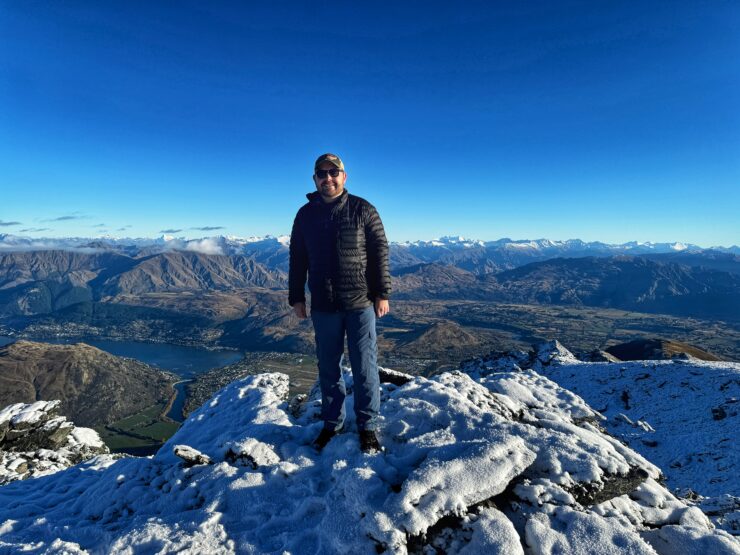 Man stands on top of mountain with snow under feet and distant mountain range behind him