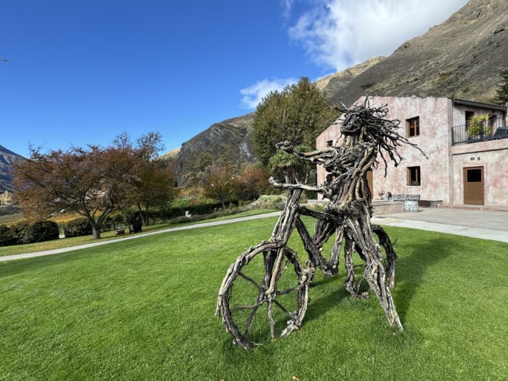 A straw figure on a bicycle sits in front of a farmhouse against a blue sky and background of hills