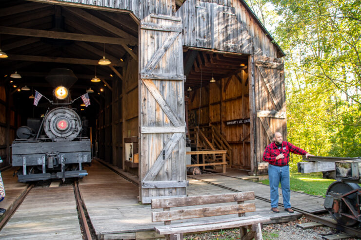 Man in a red plaid shirt and blue jeans in front of an historic train depot.
