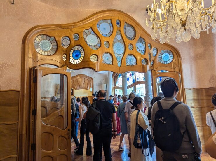 Interior of building with wide wooden doorway with curved elements. Above the door are many round stained glass insets. The area is crowded with tourists. 