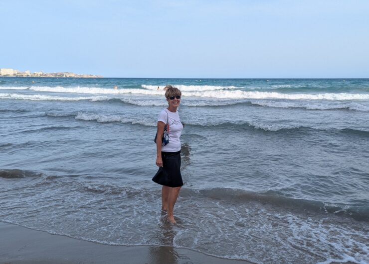 A woman in a black skirt and white T-shirt standing barefoot in ankle deep water in the Mediterraneum Sea. 