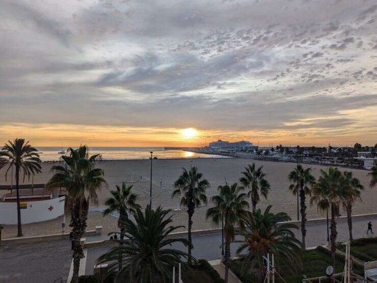 Beach sunset with palm trees fronting a wide sandy beach and a large cruise ship at a distant dock. 