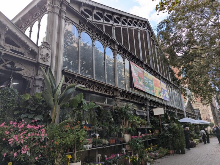 Large iron and glass neighborhood market with peaked tile roof and buckets of flowers and plants lining the front. 