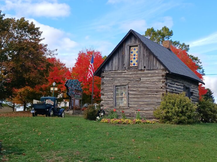 Small, old long cabin-like building next to a 1930s truck, surrounded by bright orange-leafed trees