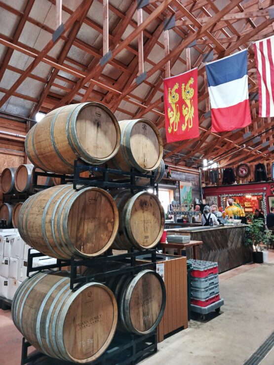 Inside a high-ceilinged barn, flags strung across the ceiling, with stacks of 300-liter barrels 3 high and 2 across next to a bar