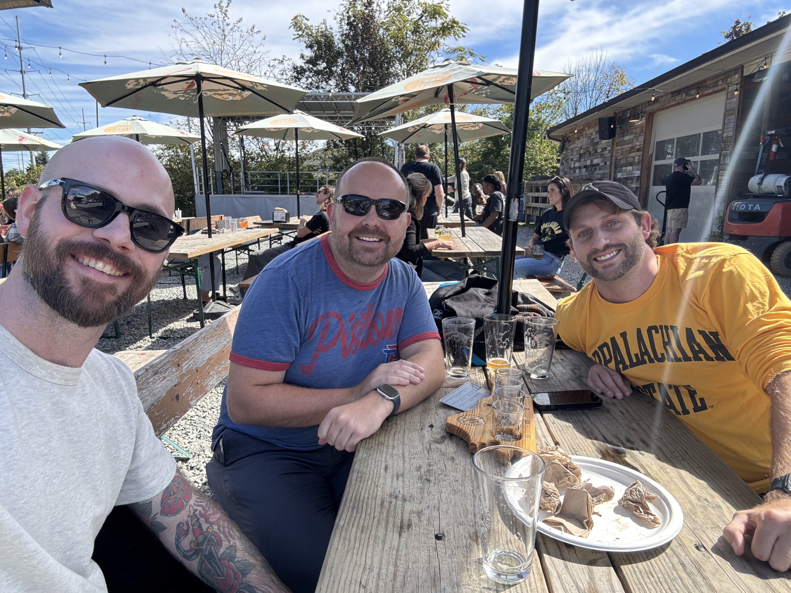 Three men in sunglasses sit in a beergarden with table and pints of beer between them