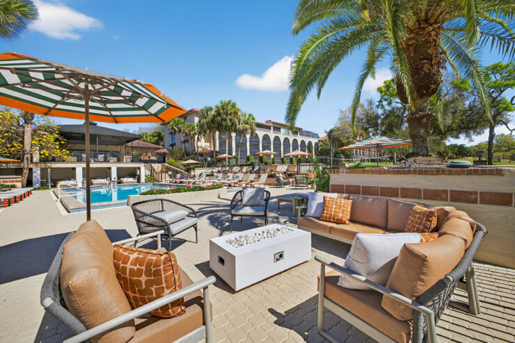 Outdoor poolside lounge area at Mission Resort + Club with cushioned seating, fire pits, umbrellas, palm trees, and resort buildings in the background.