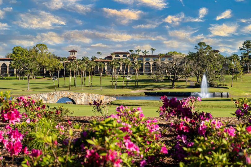Colorful flower gardens frame a view of El Campeón golf course at the Mission Resort + Club, with manicured greens, palm trees, and resort architecture under a bright blue sky.