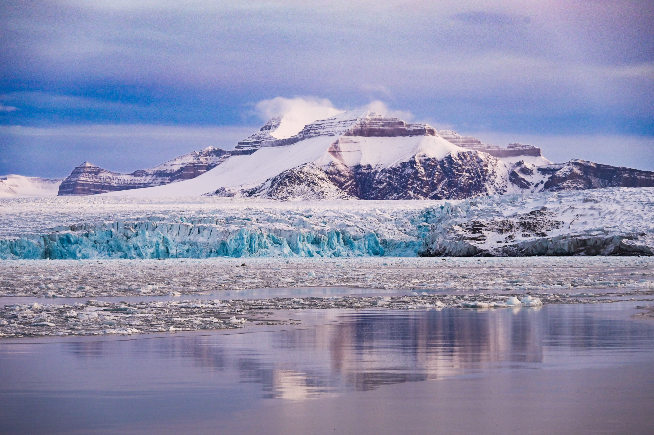 Snow-covered rocky mountains rise above a blue-tinged glacier. Both are reflected in an icy bay. The sky is a gloomy blueish-gray.