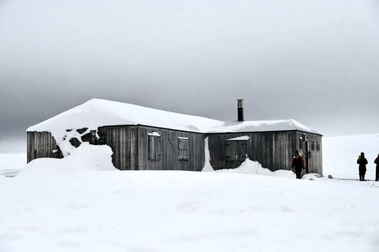An abandoned wooden building is surrounded by deep snow under a gray sky.