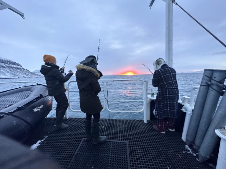 Three people are fishing off the back end of a boat. In the background is the orange setting sun reflecting on the water.