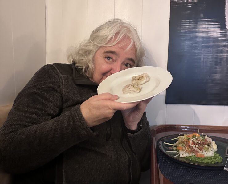 A woman with gray hair shows off a plate with two small poached fish filets. She is smiling.