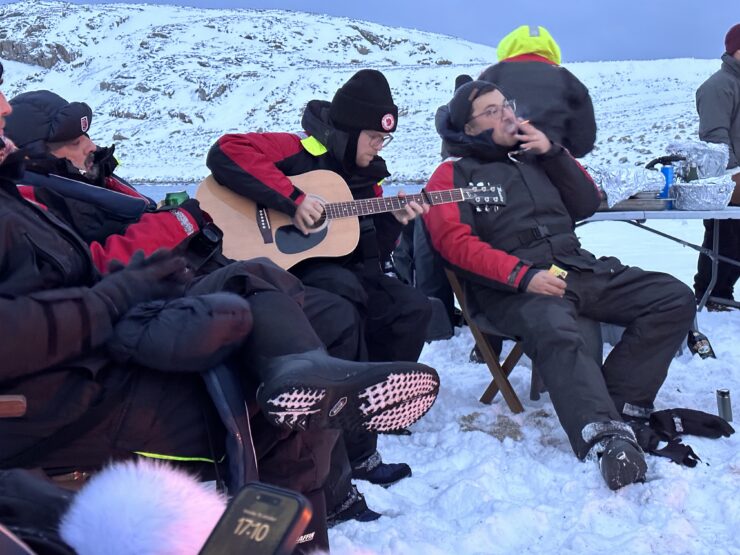 A man plays a guitar while sitting on a chair on the ice next to a glacier. There are several people sitting around him and all are wearing polar survival suits.