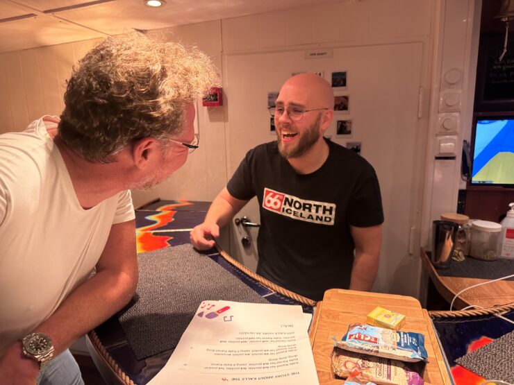 A bald, bearded man stands behind a counter covered in papers and energy bars. He is laughing with a bushy haired man on the other side of the counter.