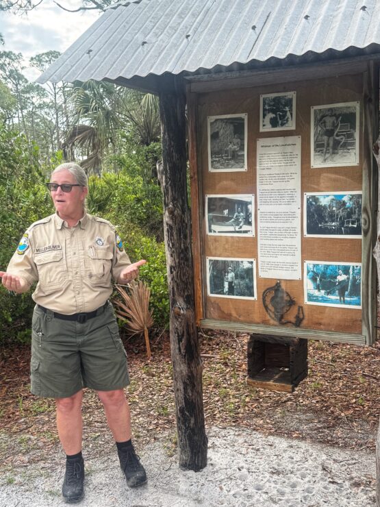 A woman in guide uniform of a beige shirt and green shorts stands in front of a populated bulletin board