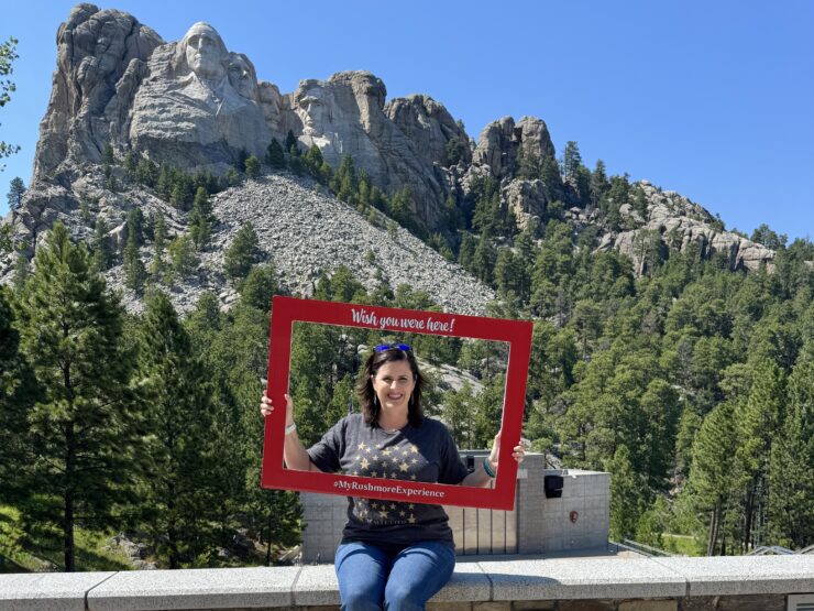 Woman wearing a grey shirt and jeans holding up a red frame in front of Mount Rushmore.