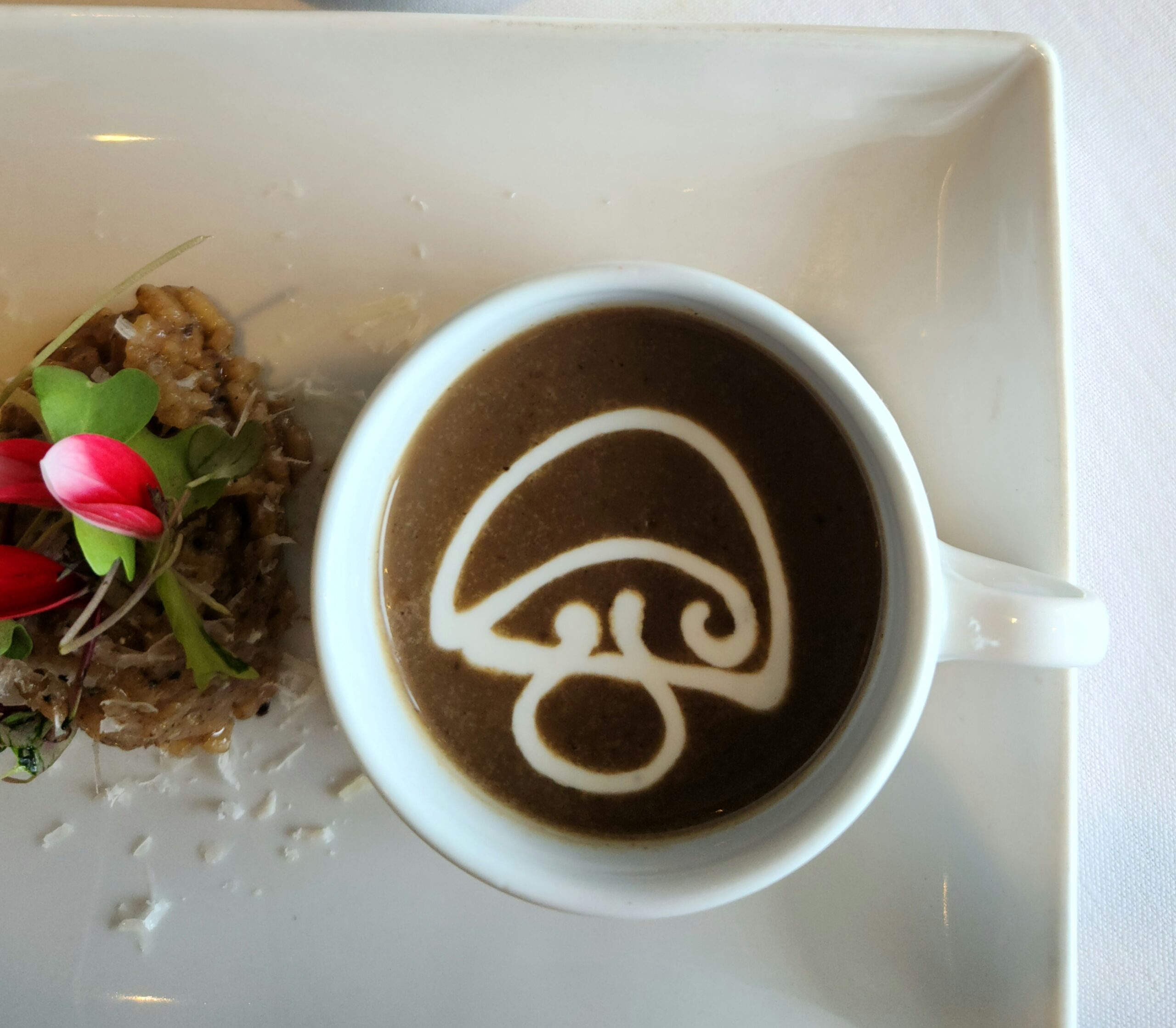 A white cup of brown mushroom soup, with a mushroom drawn in creamy white on top.