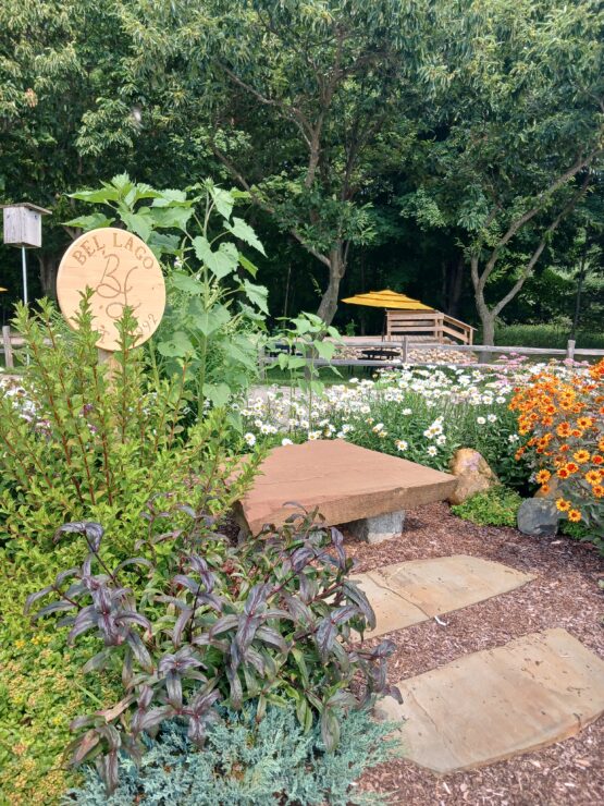 Garden overflowing with colorful flowers surrounding the Bel Lago sign in front of a wooded area